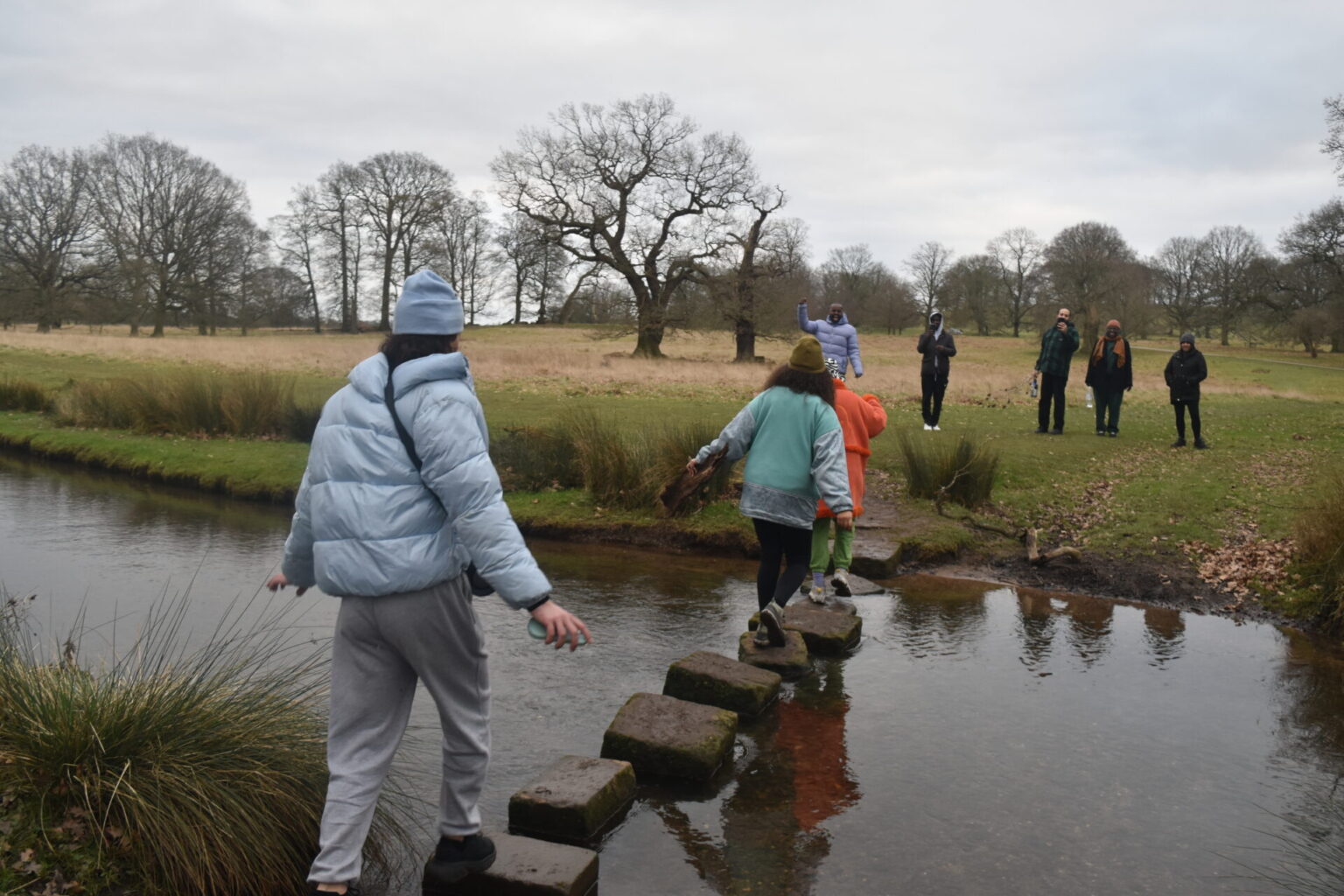 Three people walk across stones in a river. Five other people wait on the other side of the crossing.