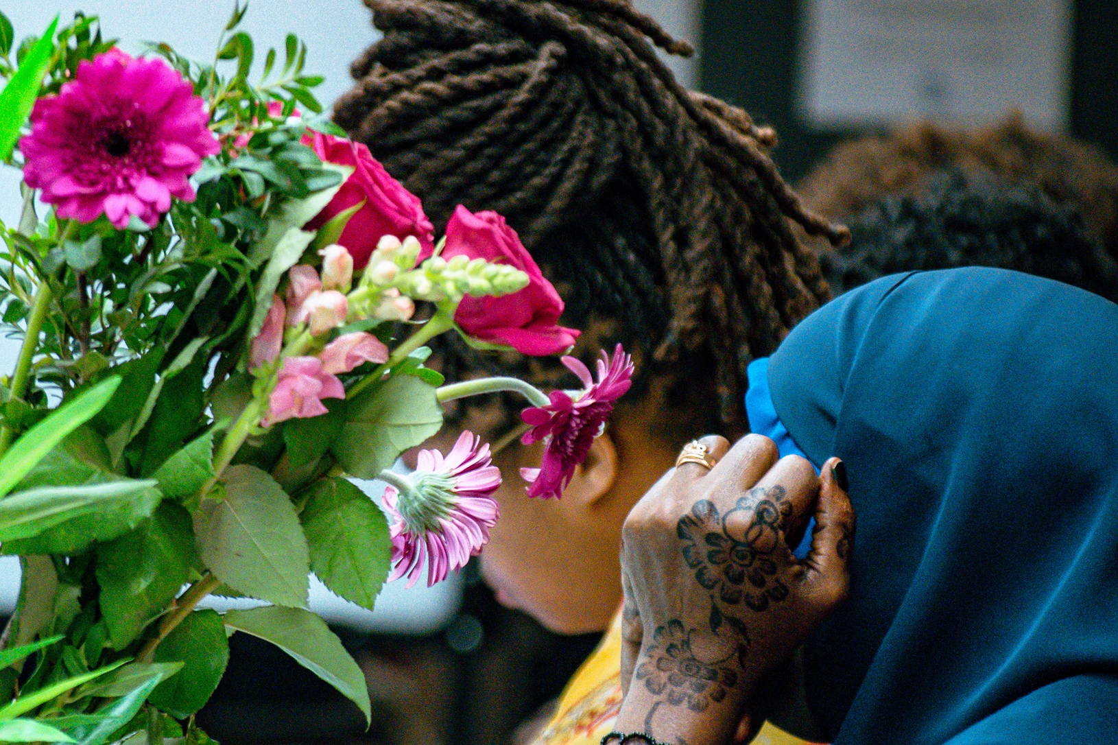 Ayuuto 2 A hijabi woman with henna on her hands next to a woman with locs - both their faces obscured. A bouquet of flowers in the foreground.