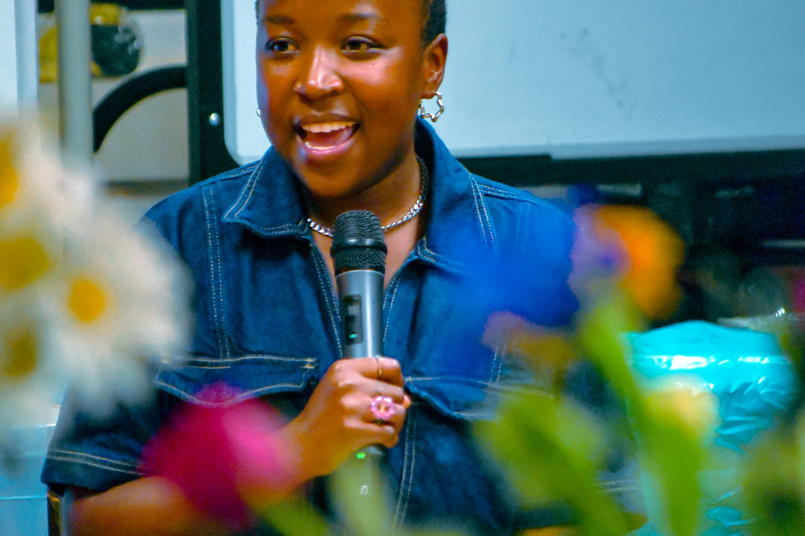 Ayuuto 5 DE founder Noni Makuyana seated, speaking into a microphone. Some out of focus plants in the foreground.