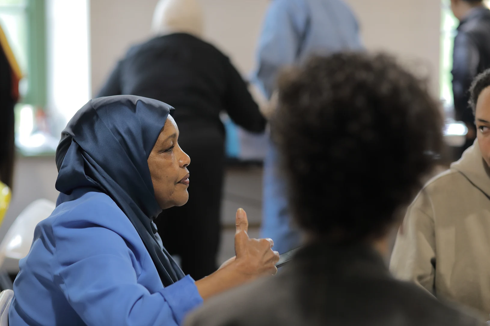 Healing in Community 3 A woman in blue gesturing with her hand. Others in the shot are out of focus and obscured